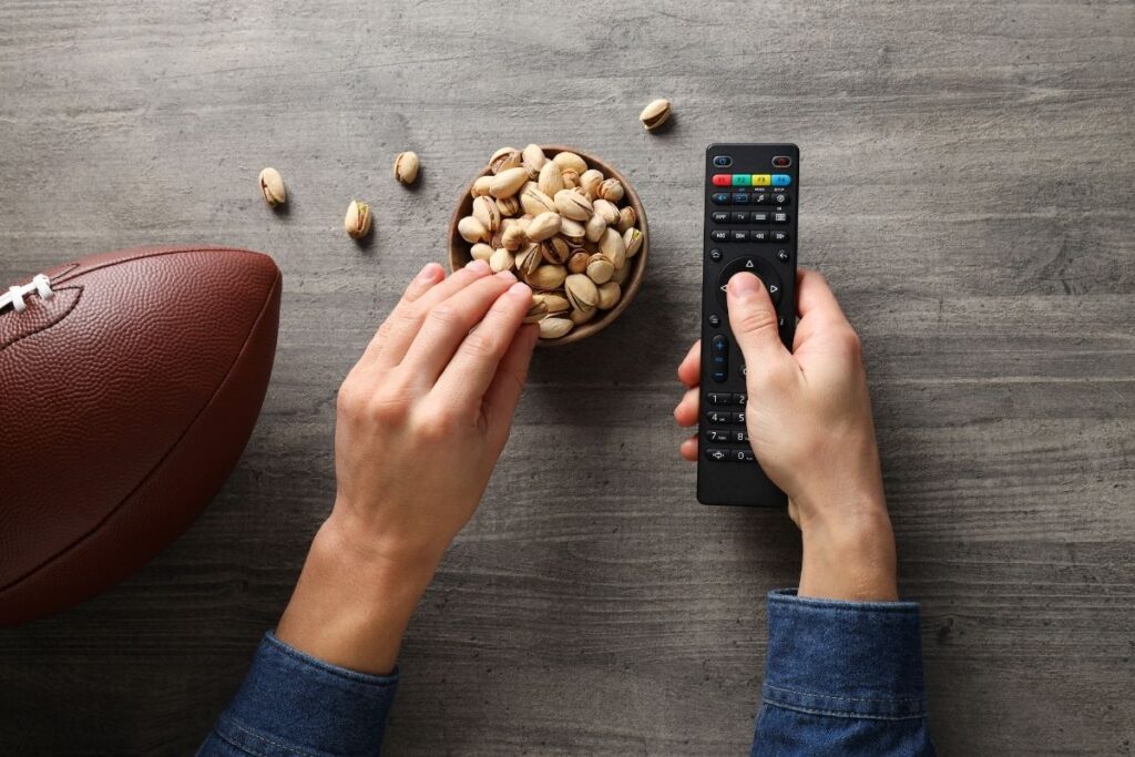 View from above of a football fan clicking the remote control and eating pistachios from a bowl with a football on the coffee table
