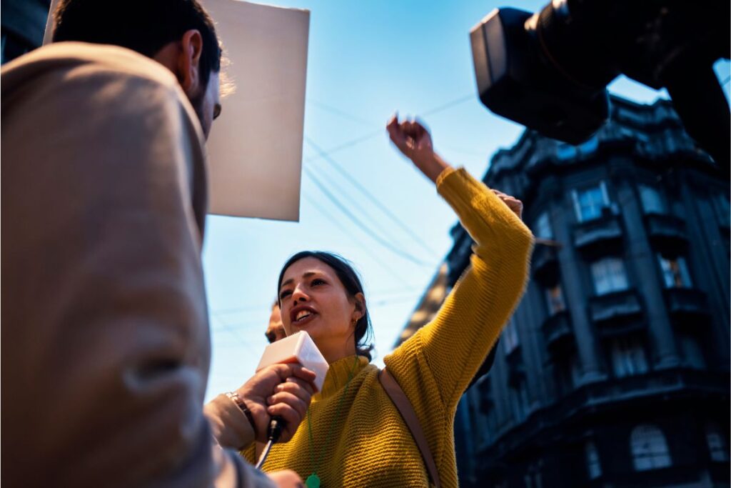 A young woman of color raises her arm emphatically while being interviewed by a TV news crew during a protest