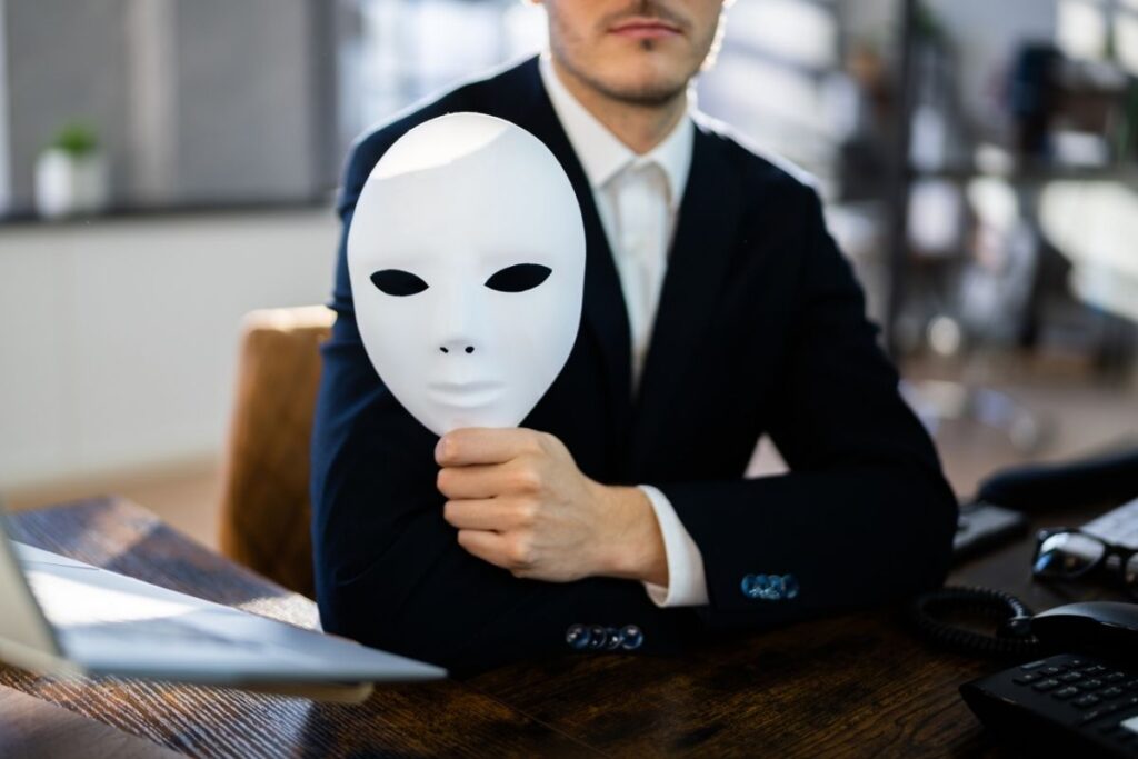 A CEO in a suit at a desk, whose face is out of frame but he is holding a plain white mask in his hand