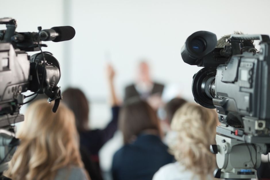 Journalists raise hands at a press conference with cameras rolling