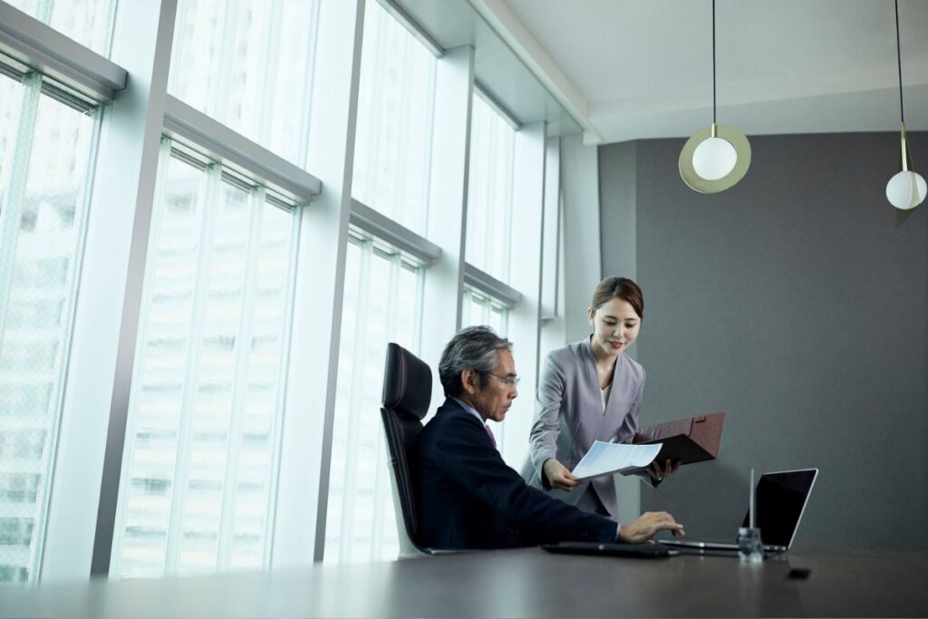 A female communications person hands a paper to an executive who is sitting by himself in a large boardroom