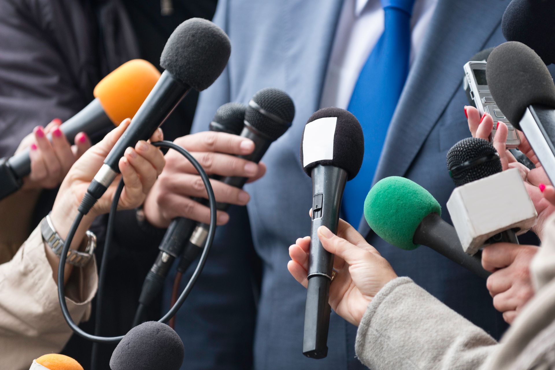 Torso of a politician in a suit, surrounded by microphones and digital recording devices.